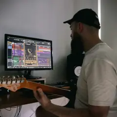 Mężczyzna z brodą i czapką z daszkiem uczy się, jak podłączyć gitarę do PC, pracując nad muzyką na komputerze.