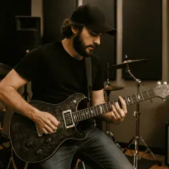 Mężczyzna gra na gitarze elektrycznej w studiu. Do gitary elektrycznej potrzeba wzmacniacza i kabla.