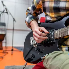 Ręce muzyka grające na czarnej gitarze elektrycznej. W tle stojaki z mikrofonami i bęben. To może być początek nauki, jak zmienić struny w gitarze elektrycznej.